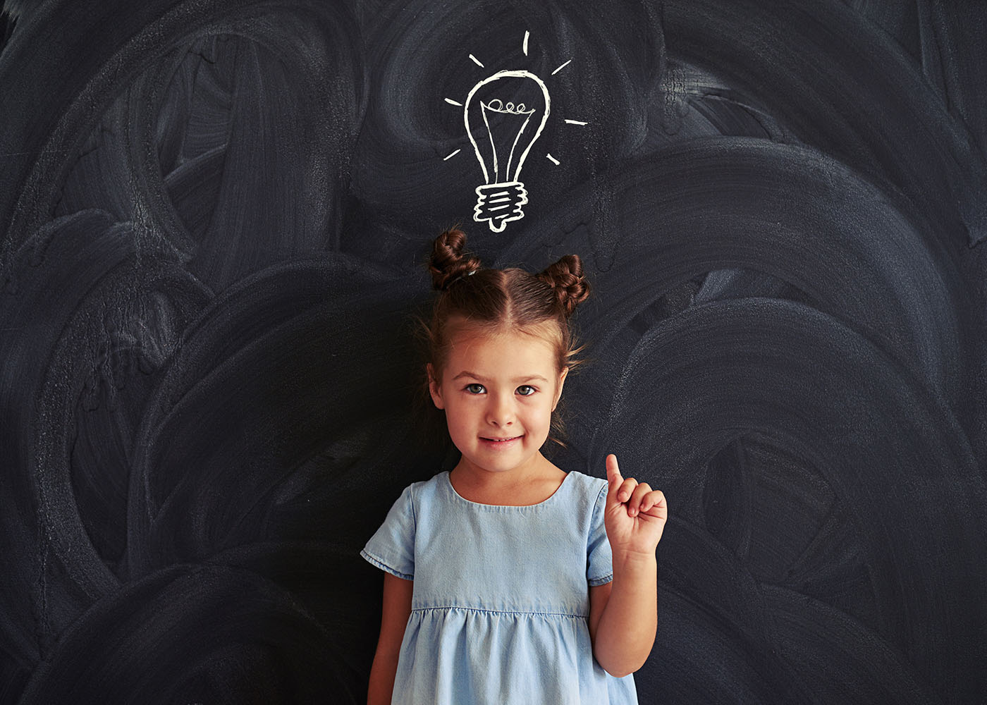 A little brown-haired girl wearing a blue dress holding up one finger standing in front of a chalkboard.  On the chalkboard behind her and above her head is the chalk drawing of a lightbulb, as if she has a bright idea.