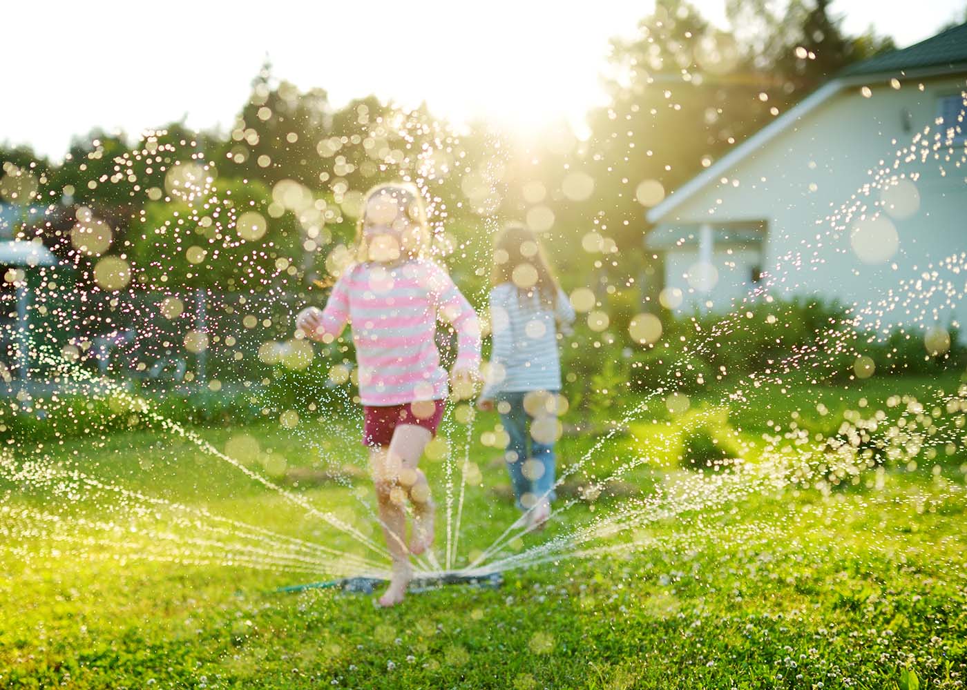 Two little girls running through a sprinkler in their backyard on a hot summer day rather than watching a TV screen