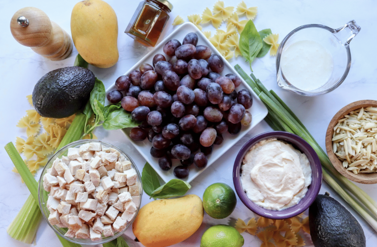 Ingredients needed to make mango chicken pasta salad spread out on a white marble countertop.