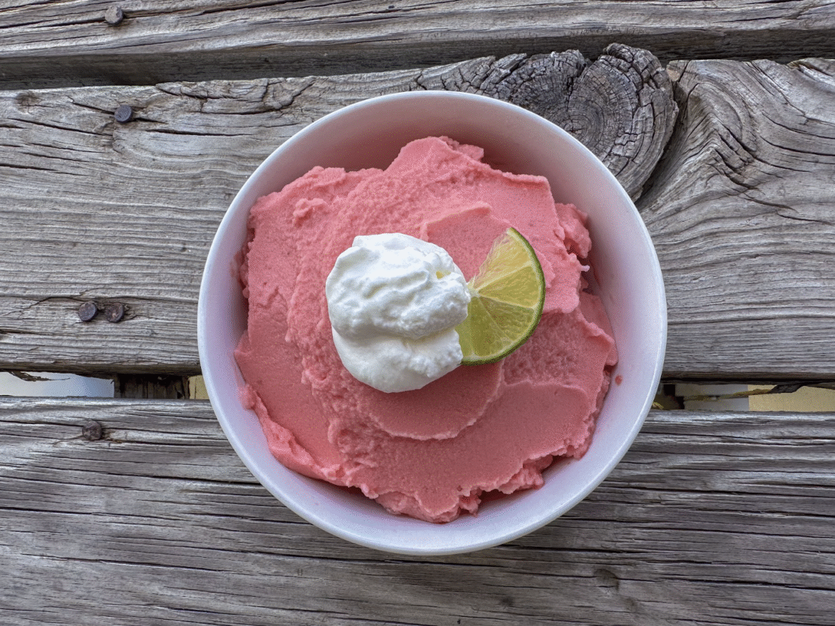 Fresh watermelon sorbet in a white bowl on a wooden picnic table, topped with a dollop of whipped cream and a slice of lime. 