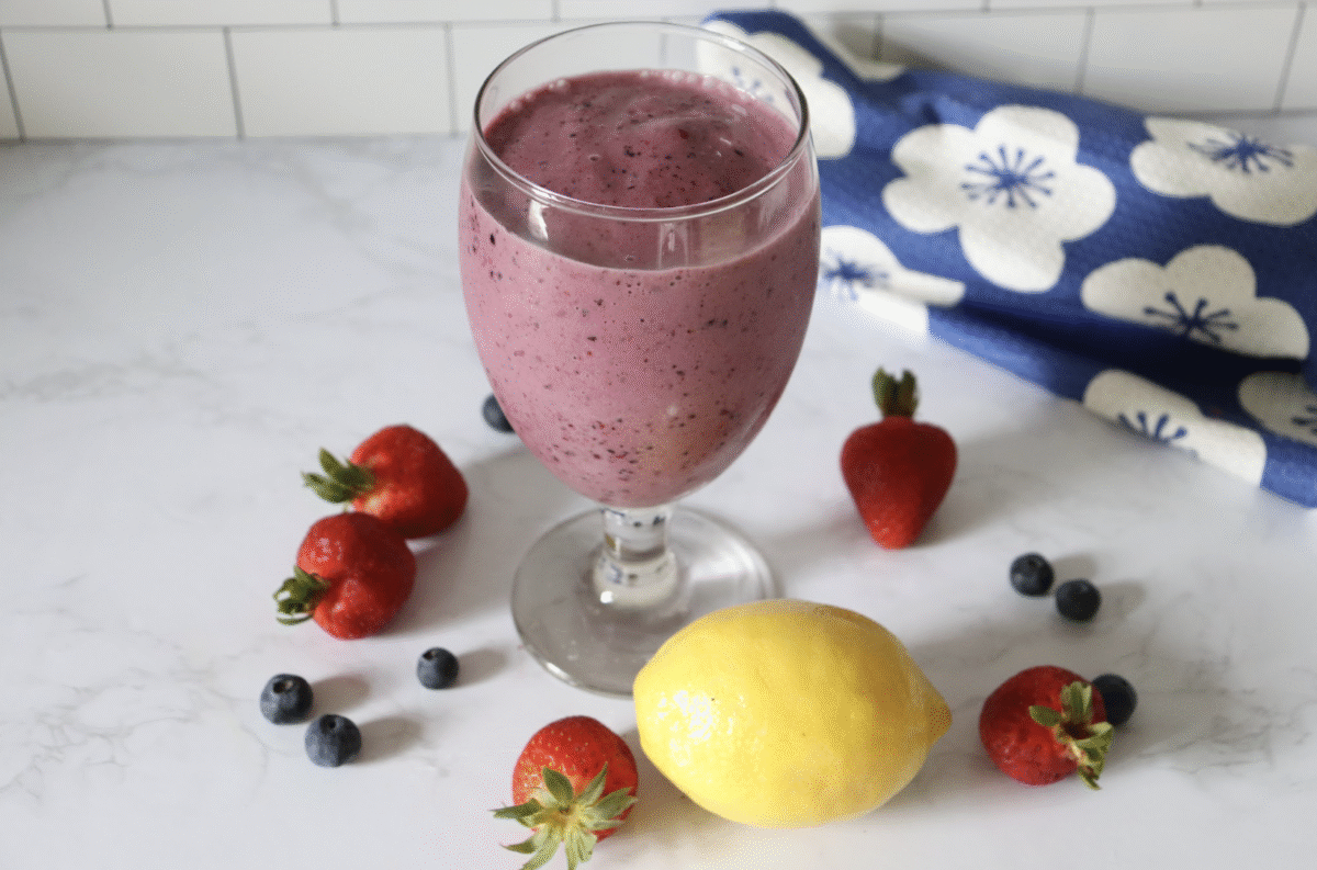 A stemmed glass filled with lemon berry power smoothie on a marble countertop with strawberries, blueberries and a lemon. 