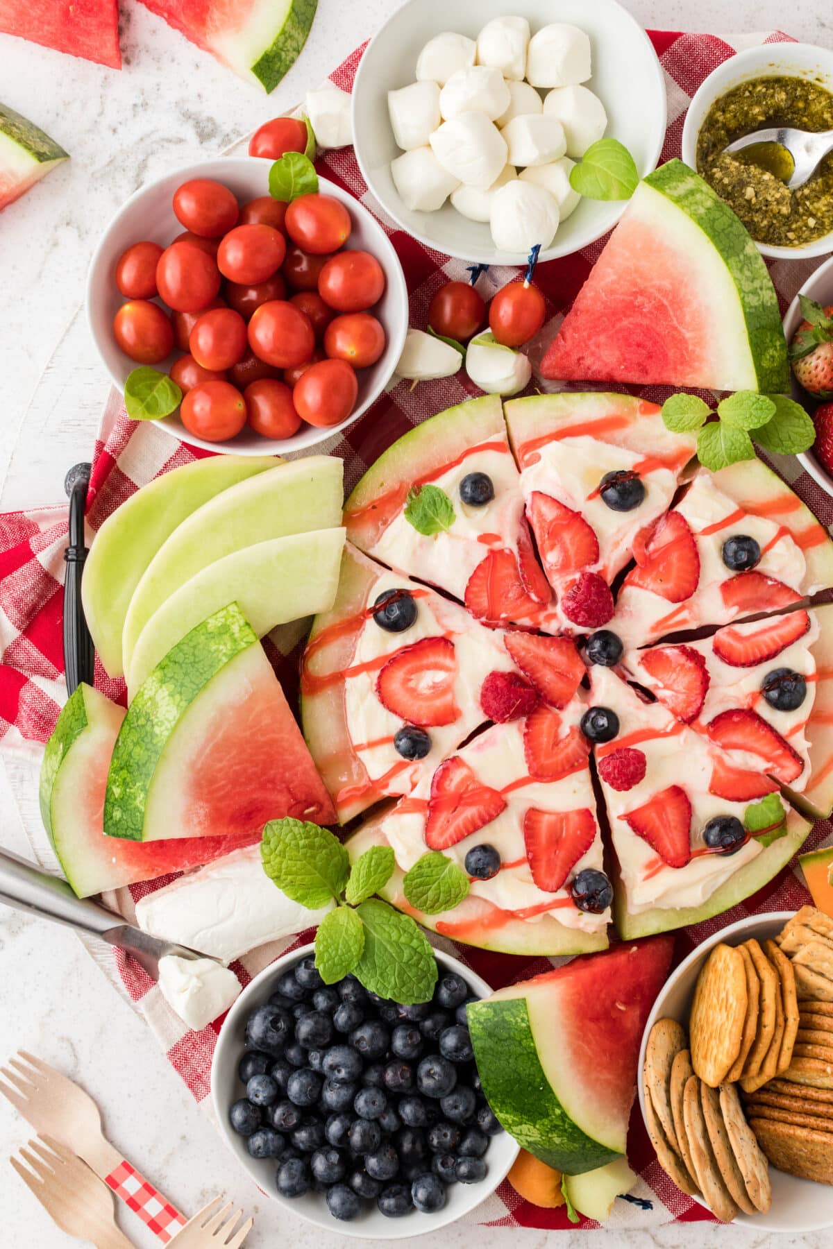 A watermelon pizza topped with whipped cream and fresh berries on a brightly colored charcuterie board.