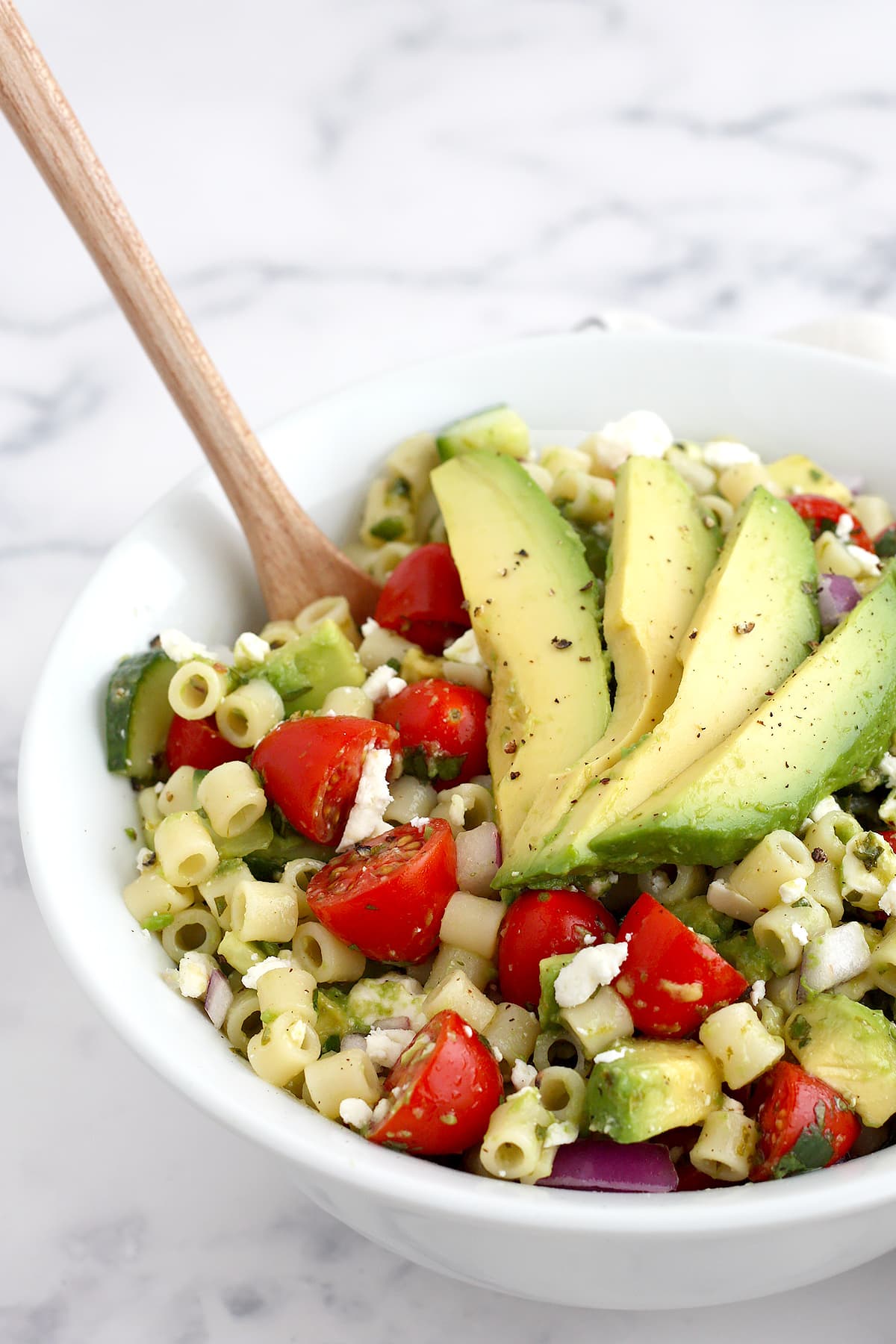 Pasta, tomatoes, cucumbers and avocado in a bowl with a wooden serving spoon. 