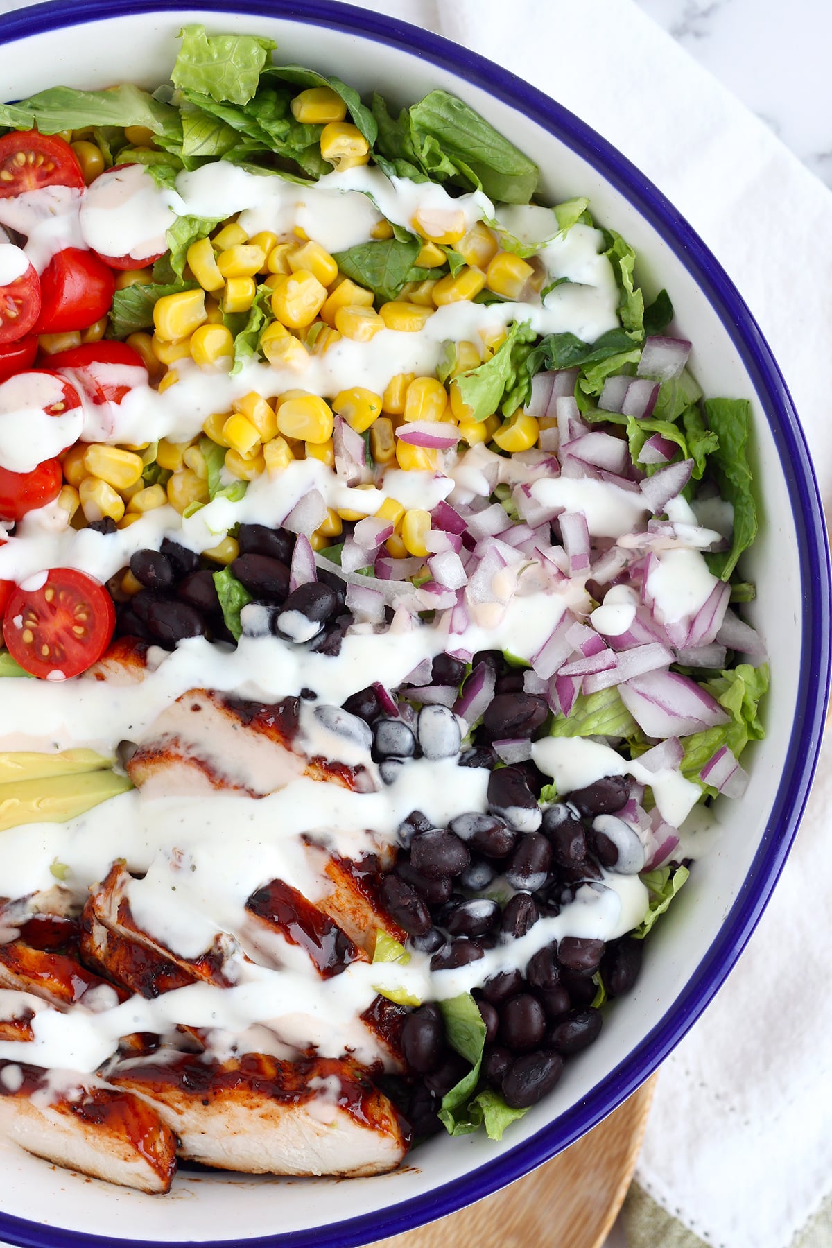 BBQ Chicken salad with grilled chicken, black beans, avocado, tomatoes, and drizzled with a bbq ranch dressing in a white and blue serving bowl. 