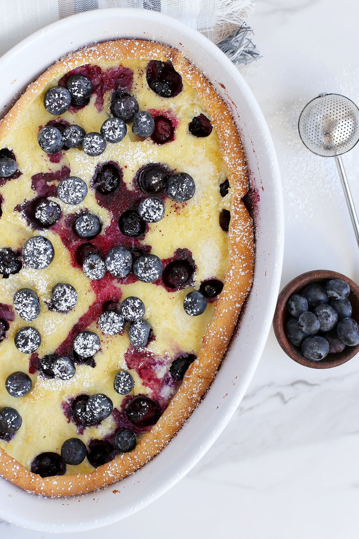 A dutch baby pancake topped with fresh blueberries and baked in a white ceramic baking dish. 