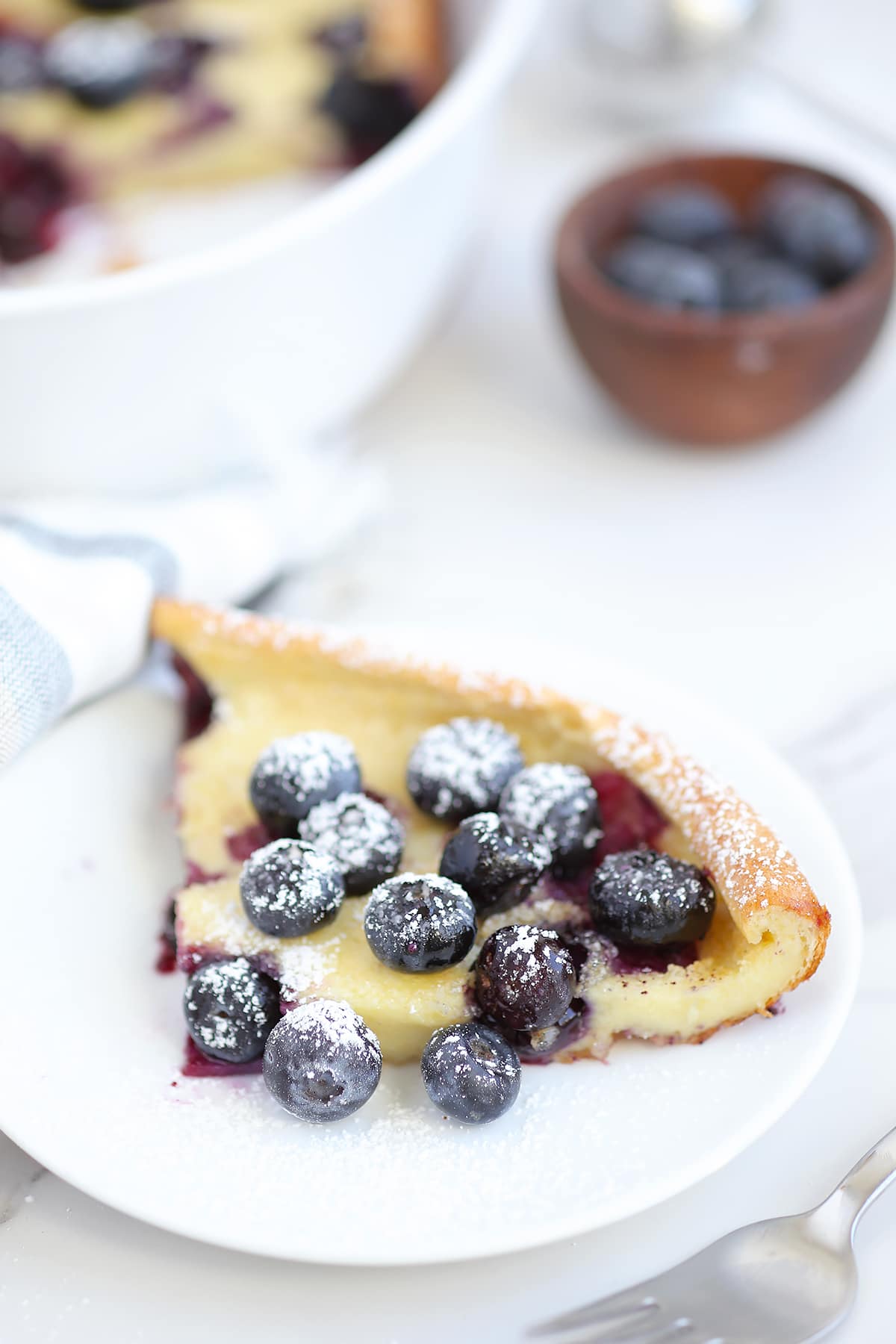 A slice of blueberry dutch baby on a white serving plate dusted with powdered sugar. 