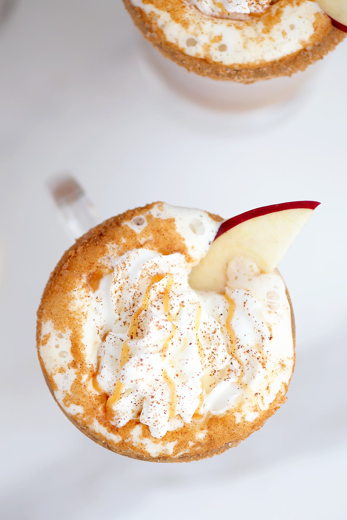 An overhead shot of a mug of cider topped with whipped cream, ground nutmeg, a caramel drizzle, and an apple slice. 