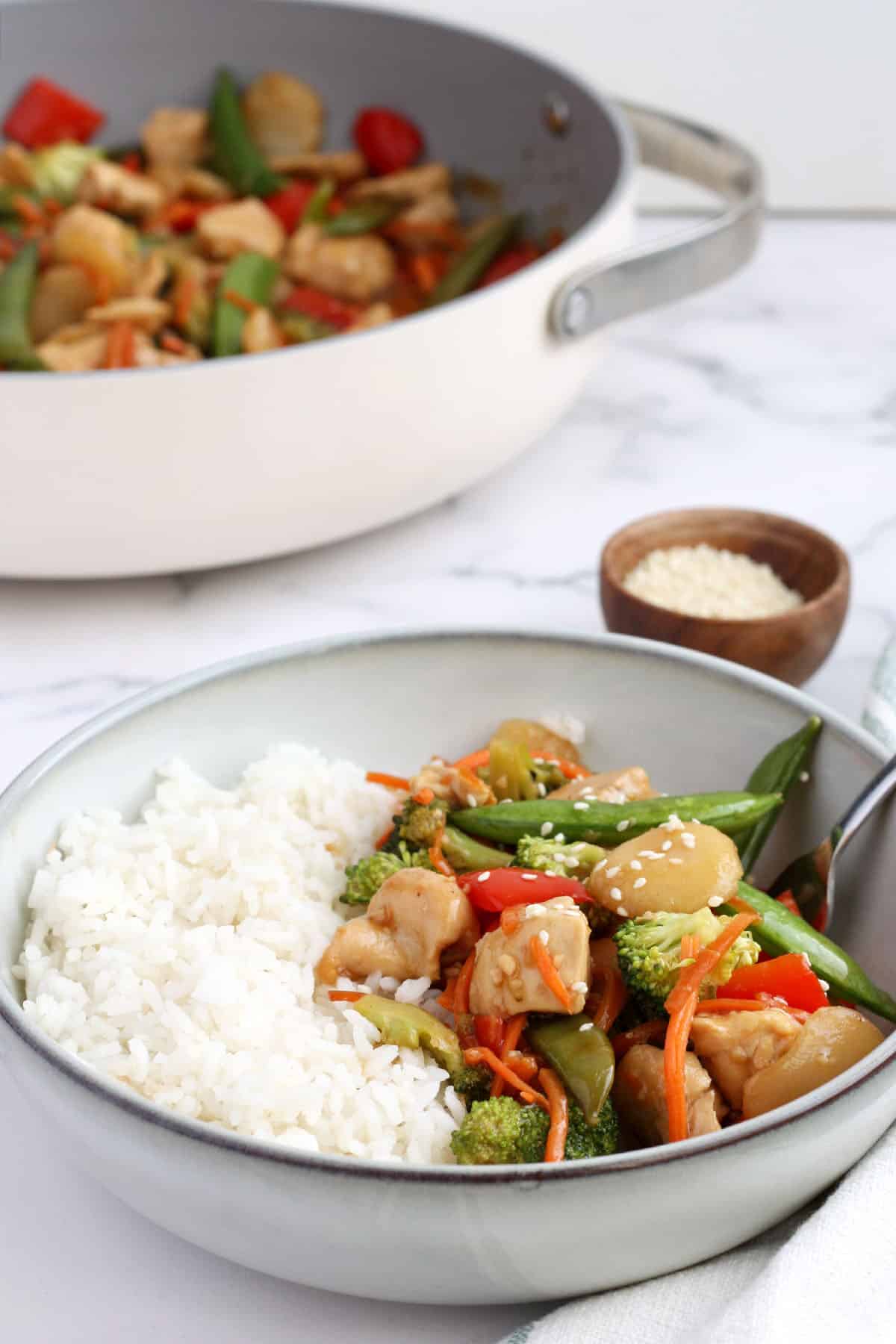 a large serving bowl with rice, chicken and veggies next to a small wooden bowl of sesame seeds