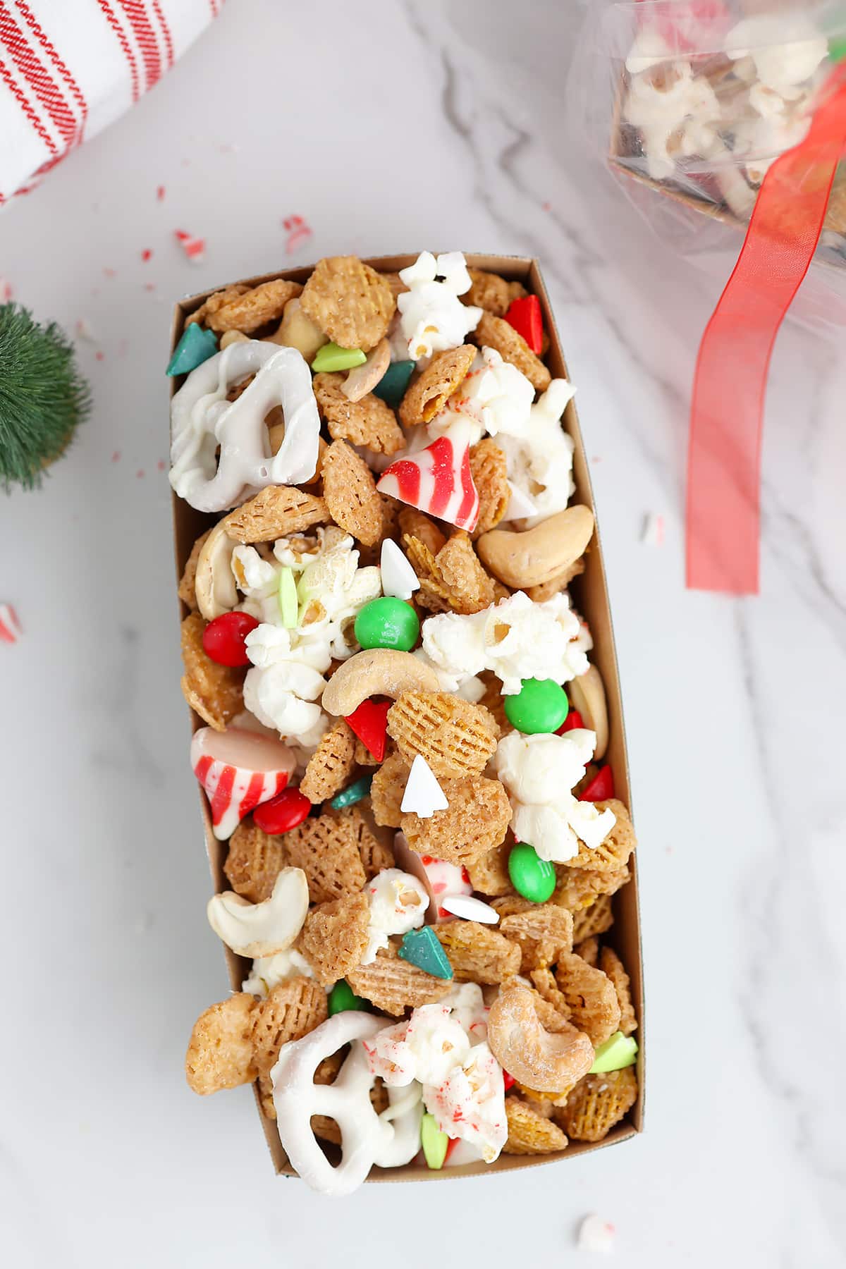 Chocolate covered pretzels, cashews, chex mix, popcorn, and candies tossed together in a white serving bowl with a large spoon. 