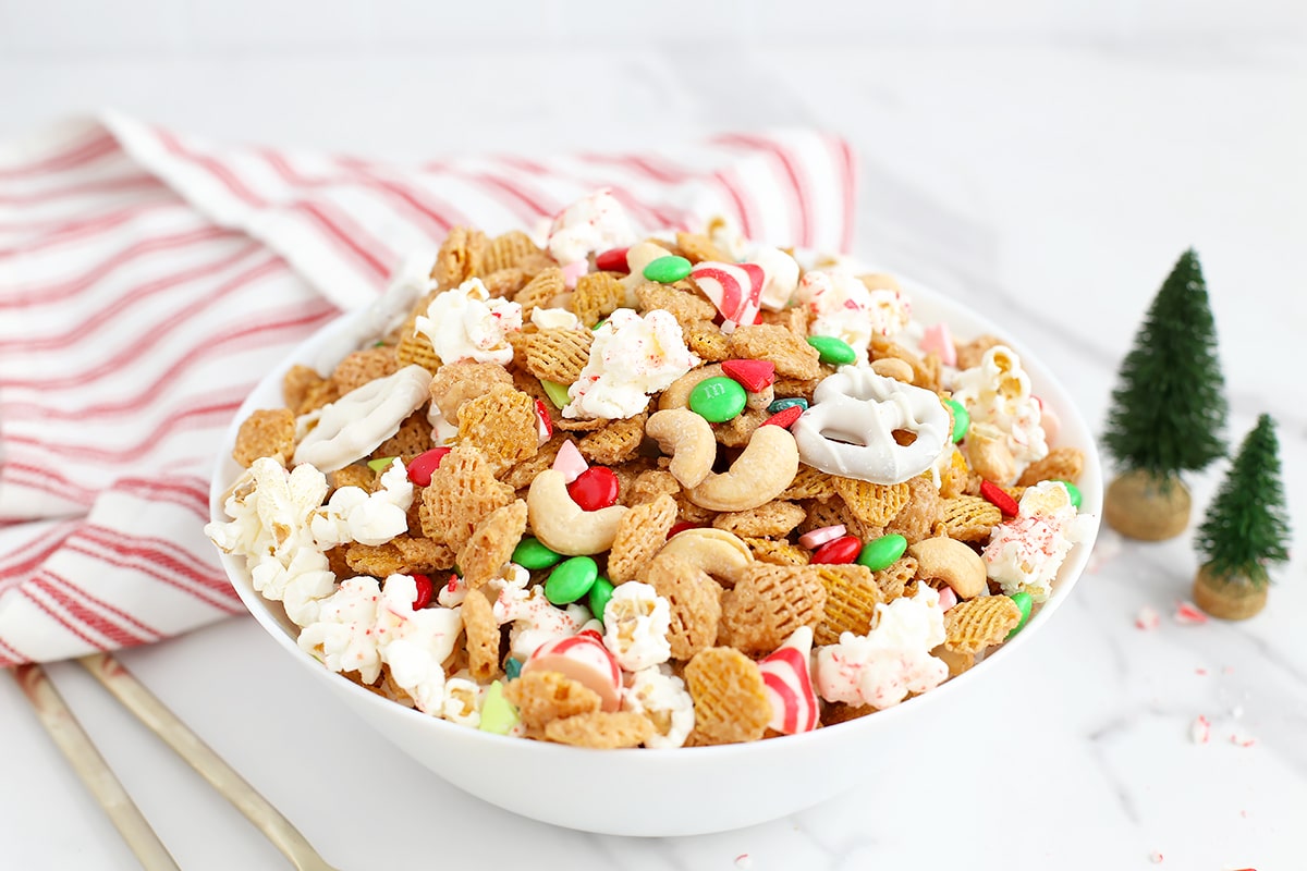 A bowl filled with holiday snack mix with a red and white striped linen and mini christmas trees in the background. 