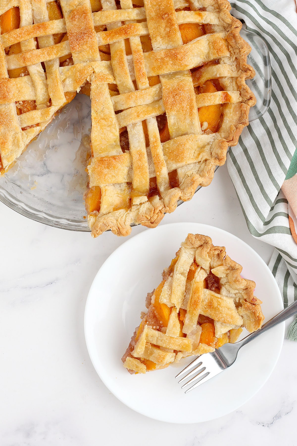 Homemade peach pie with a slice cut out and placed on a white serving plate with a striped peach linen in the background. 