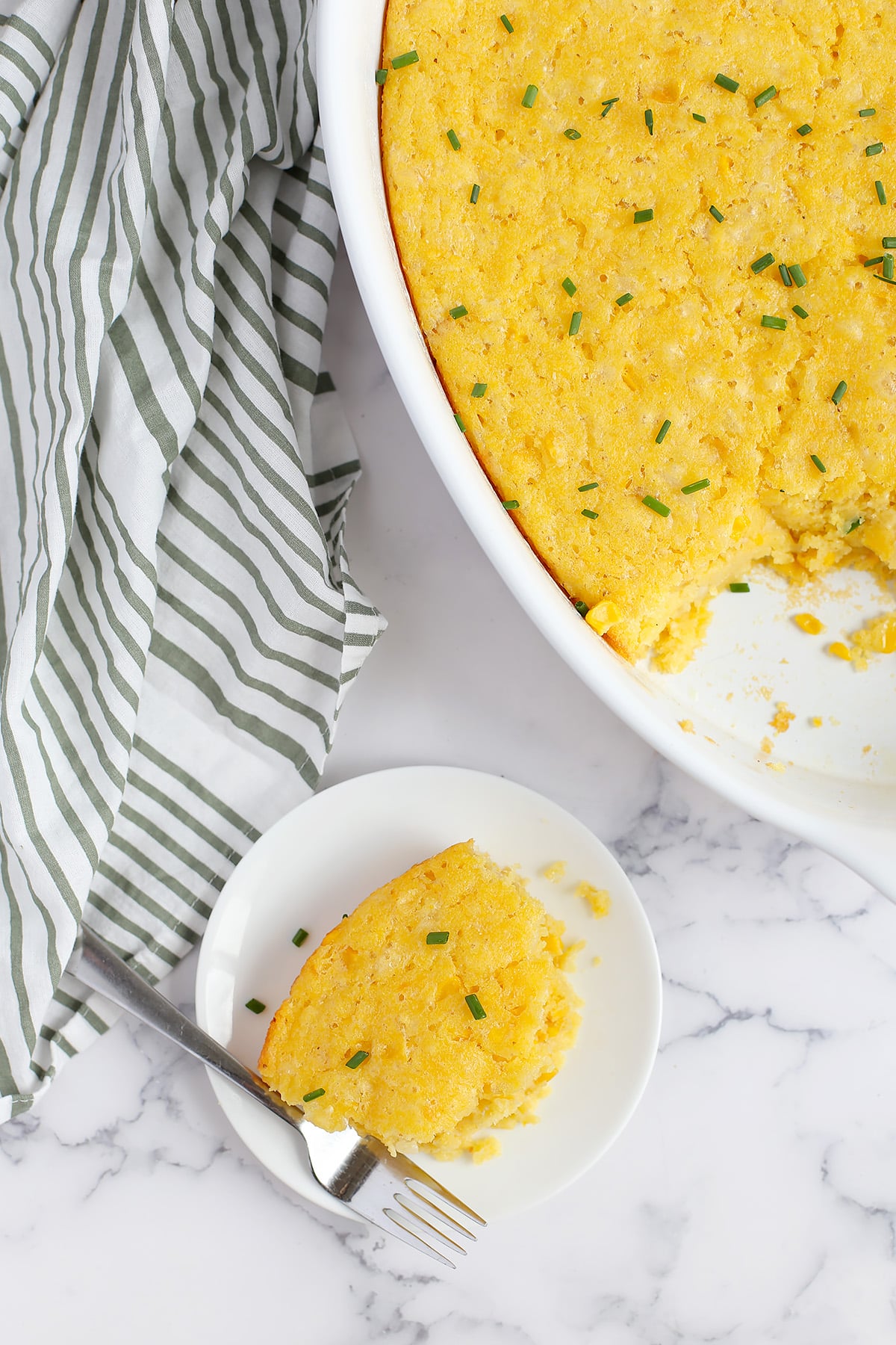An overheat shot of cornbread casserole scooped out of the baking dish and put on a serving plate with a striped linen in the background. 