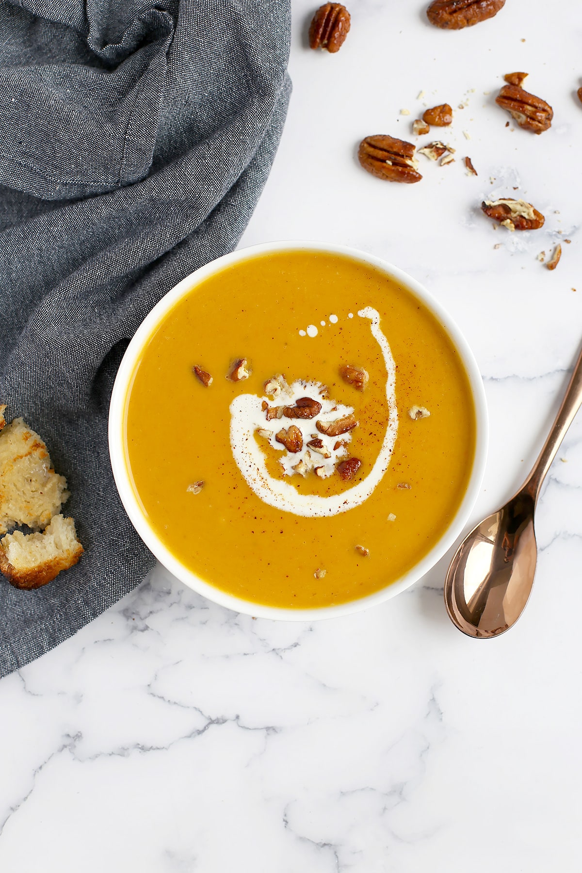 Creamy butternut squash soup in a white serving bowl, with a blue linen and a chunk of crusty bread on the side. 