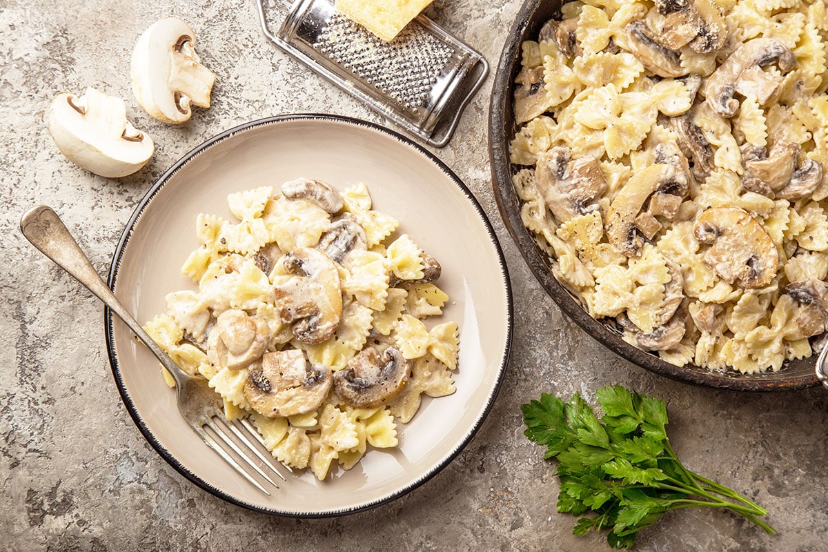 bowtie pasta with mushrooms and a creamy sauce in a cast iron skillet with parmesan cheese, parsley and mushrooms in the background.  Plate of mushroom pasta with a fork on the plate. 