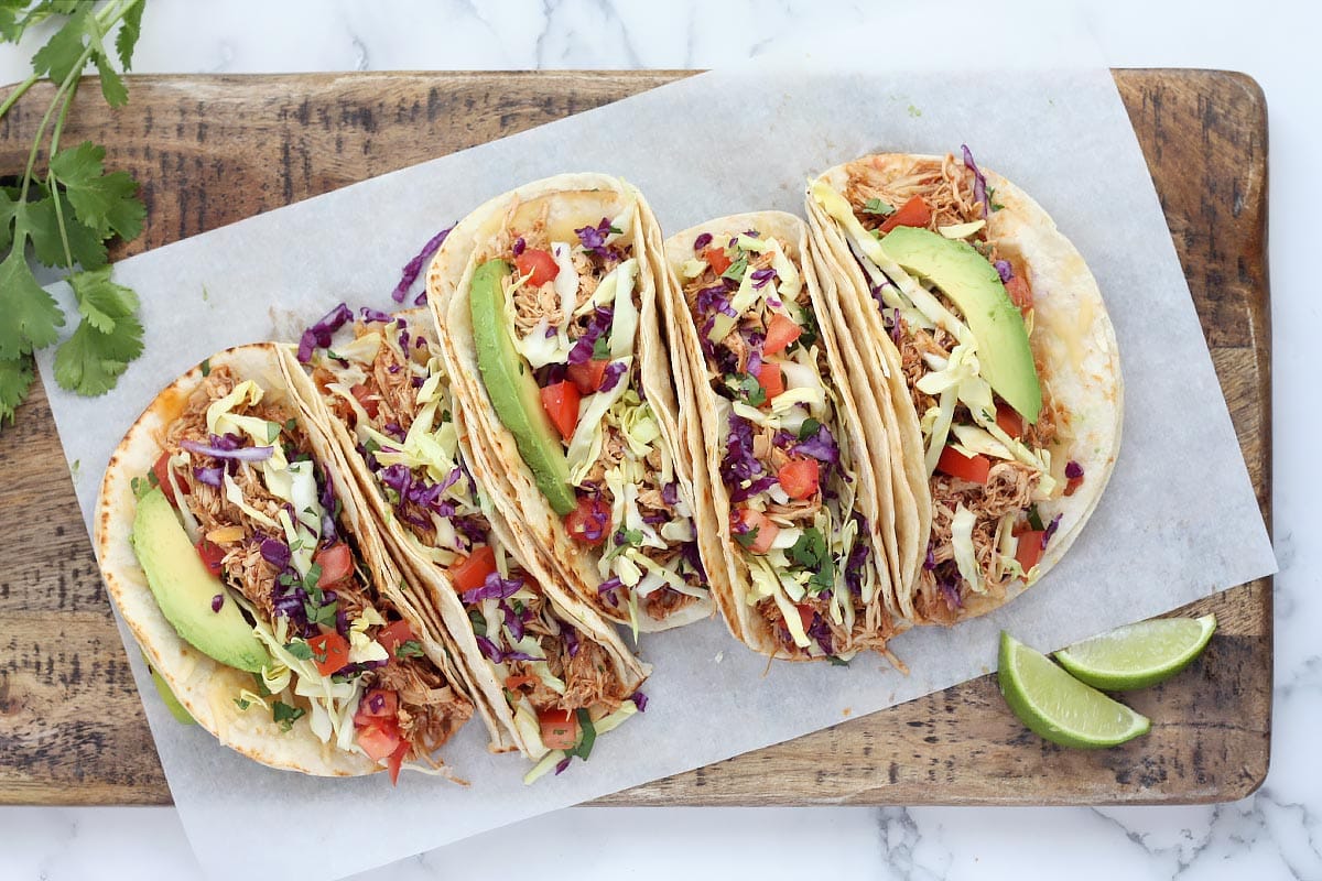 Shredded chicken tacos topped with cabbage, avocado, and tomatoes on a wooden cutting board with fresh lime slices and cilantro.