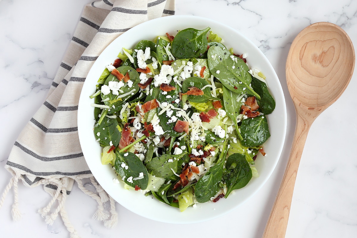 Spinach salad with crumbled bacon and feta cheese with a wooden serving spoon and striped linen.