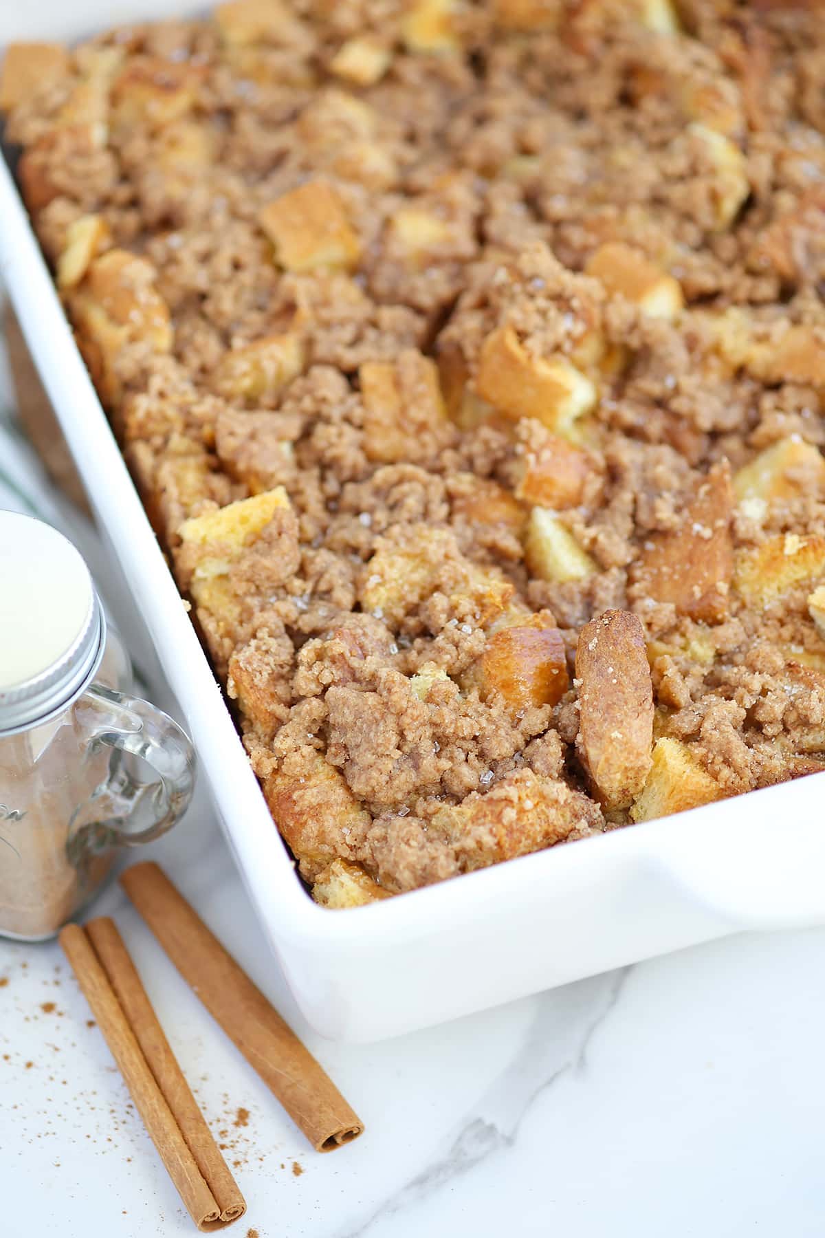 Baked French Toast in a white ceramic baking dish with cinnamon sticks and a cinnamon shaker in the background. 