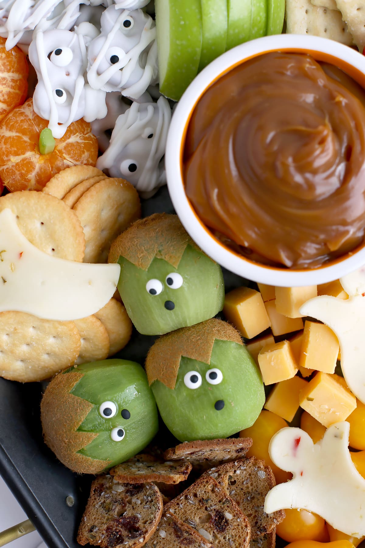 A snack tray with a bowl of caramel dip with crackers, chocolate dipped pretzels, cheese slices, and kiwi peeled to look like frankenstein monsters. 
