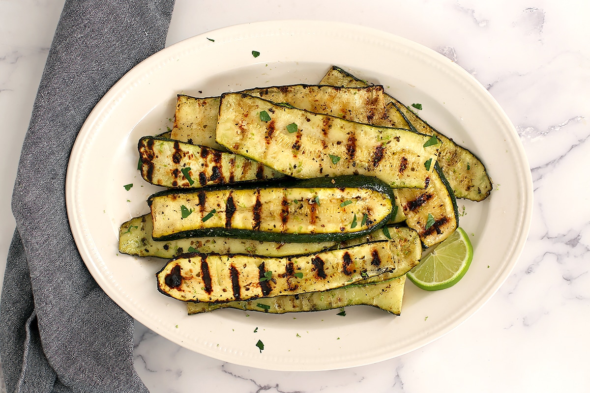 An overhead shot of zucchini slices with golden brown grill marks on an oval serving platter and a blue linen. 