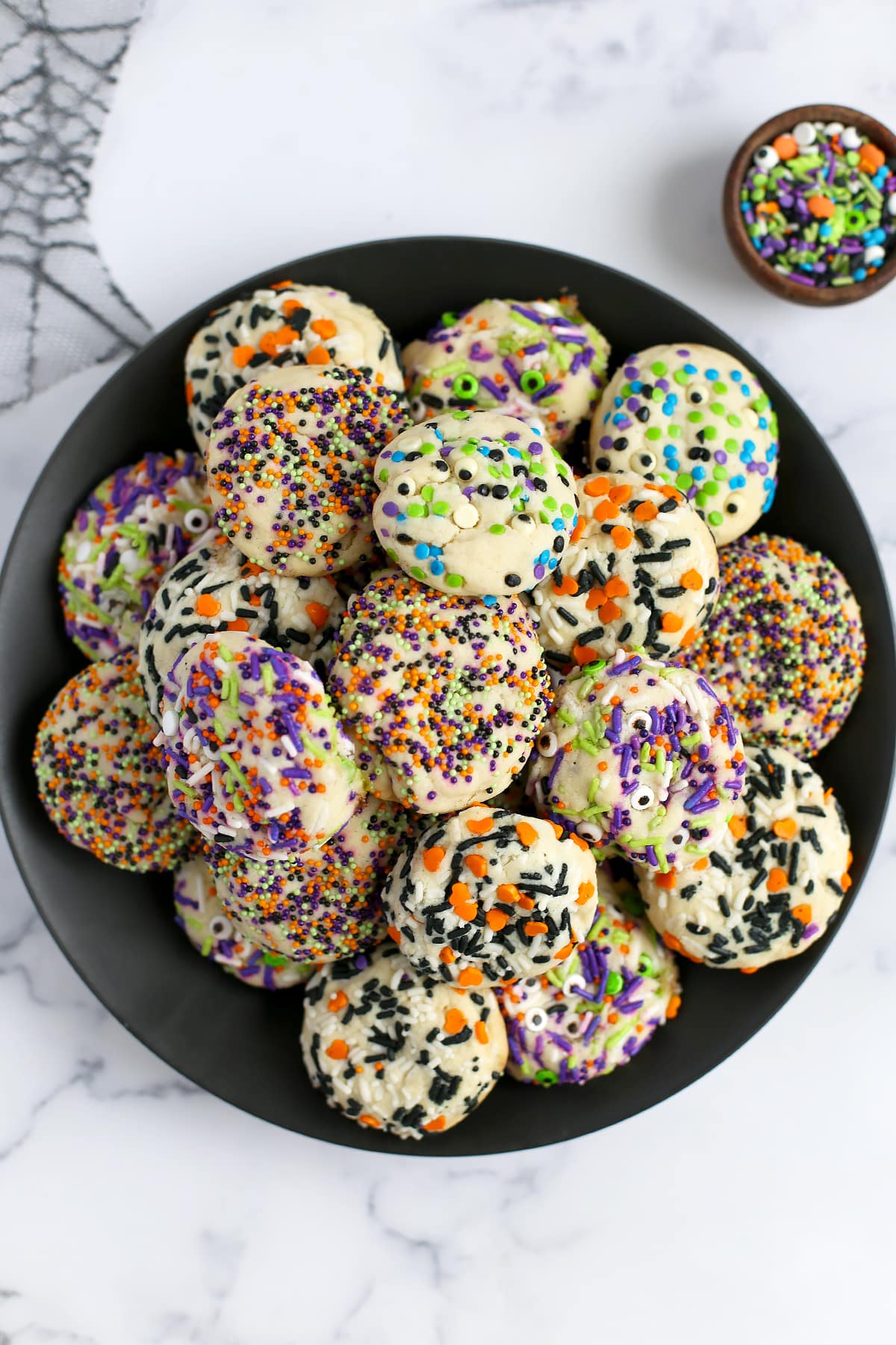 Sugar cookies rolled in Halloween sprinkles and stacked on a black plate with a spiderweb cloth in the background. 