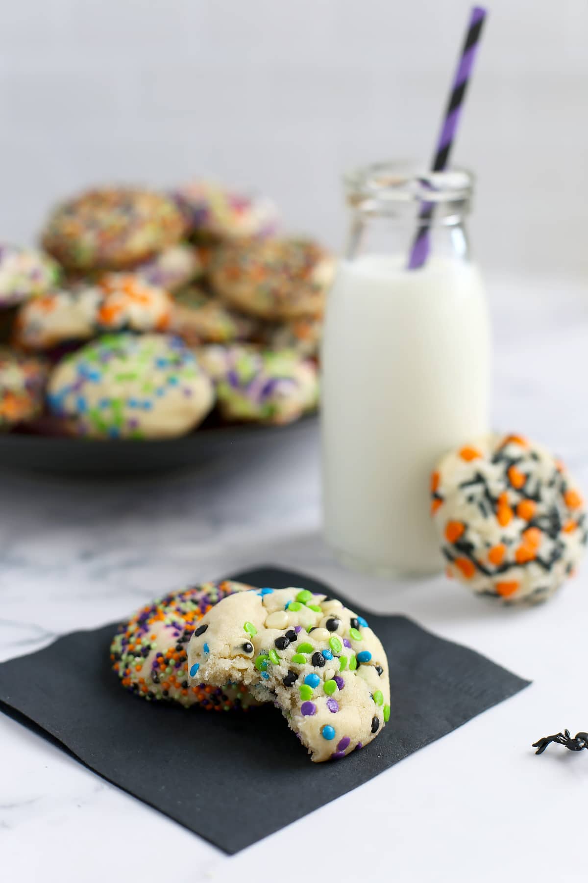 Halloween sugar cookies with a bite taken out on a black napkin with a glass bottle of milk in the background. 