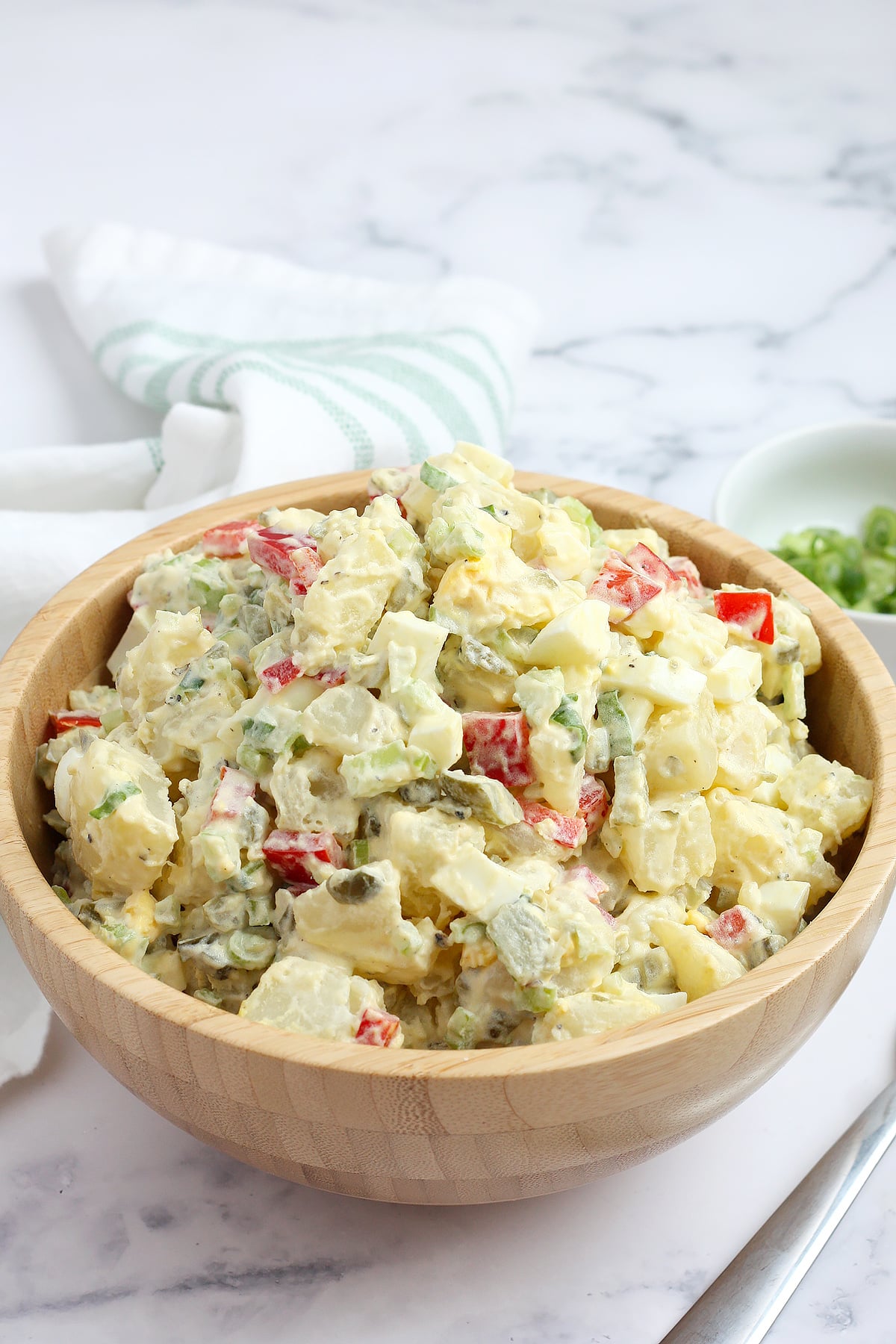 A wooden bowl full of potato salad with peppers and celery on a white table. 