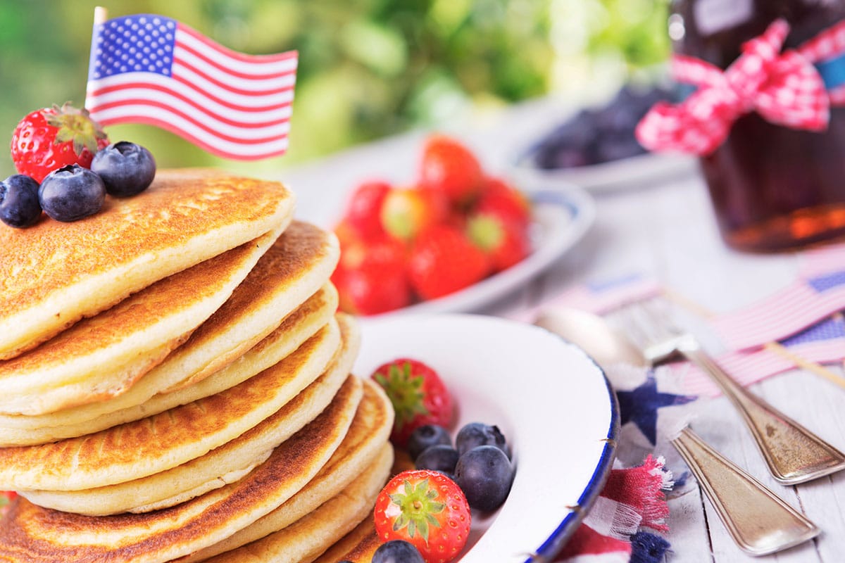 A stack of homemade whole grain pancakes with fresh berries, butter and syrup and an american flag on top
