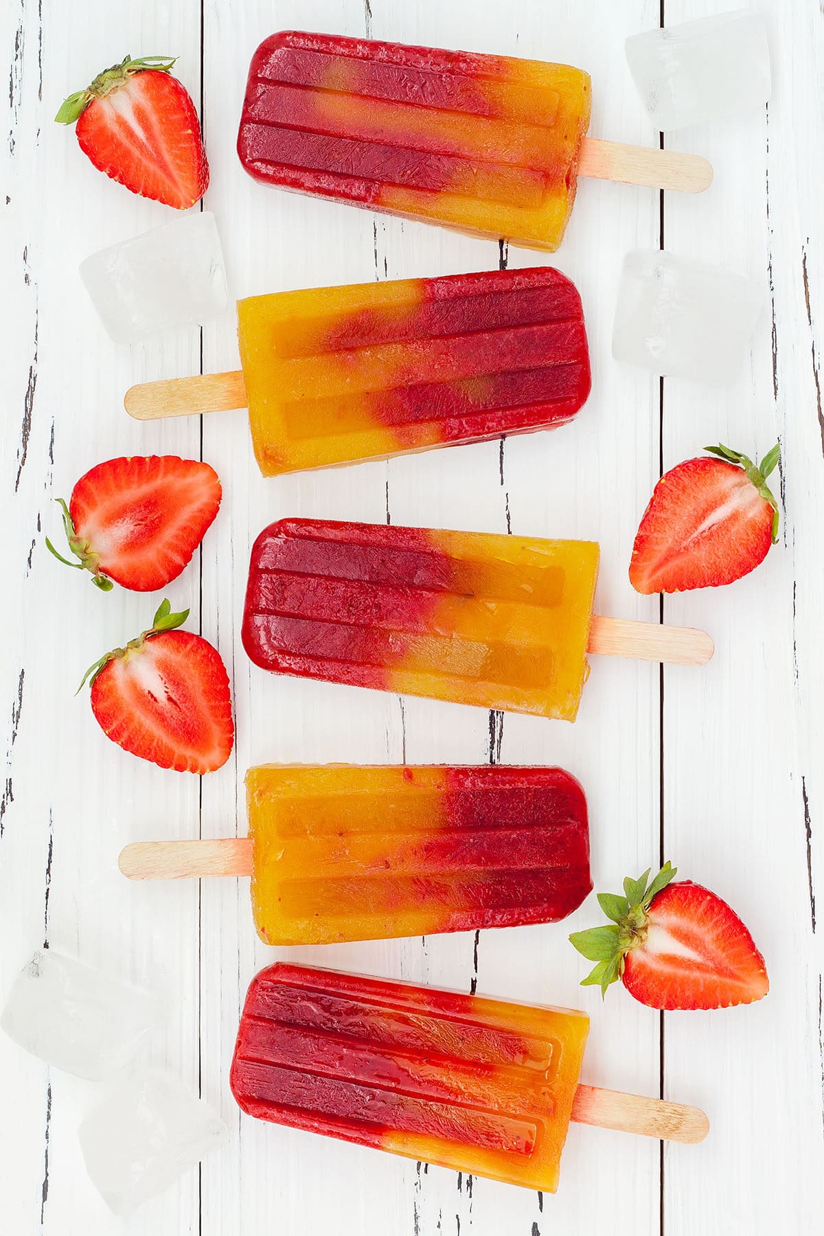 homemade strawberry mango popsicles in a line with fresh strawberries and ice cubes on a white board
