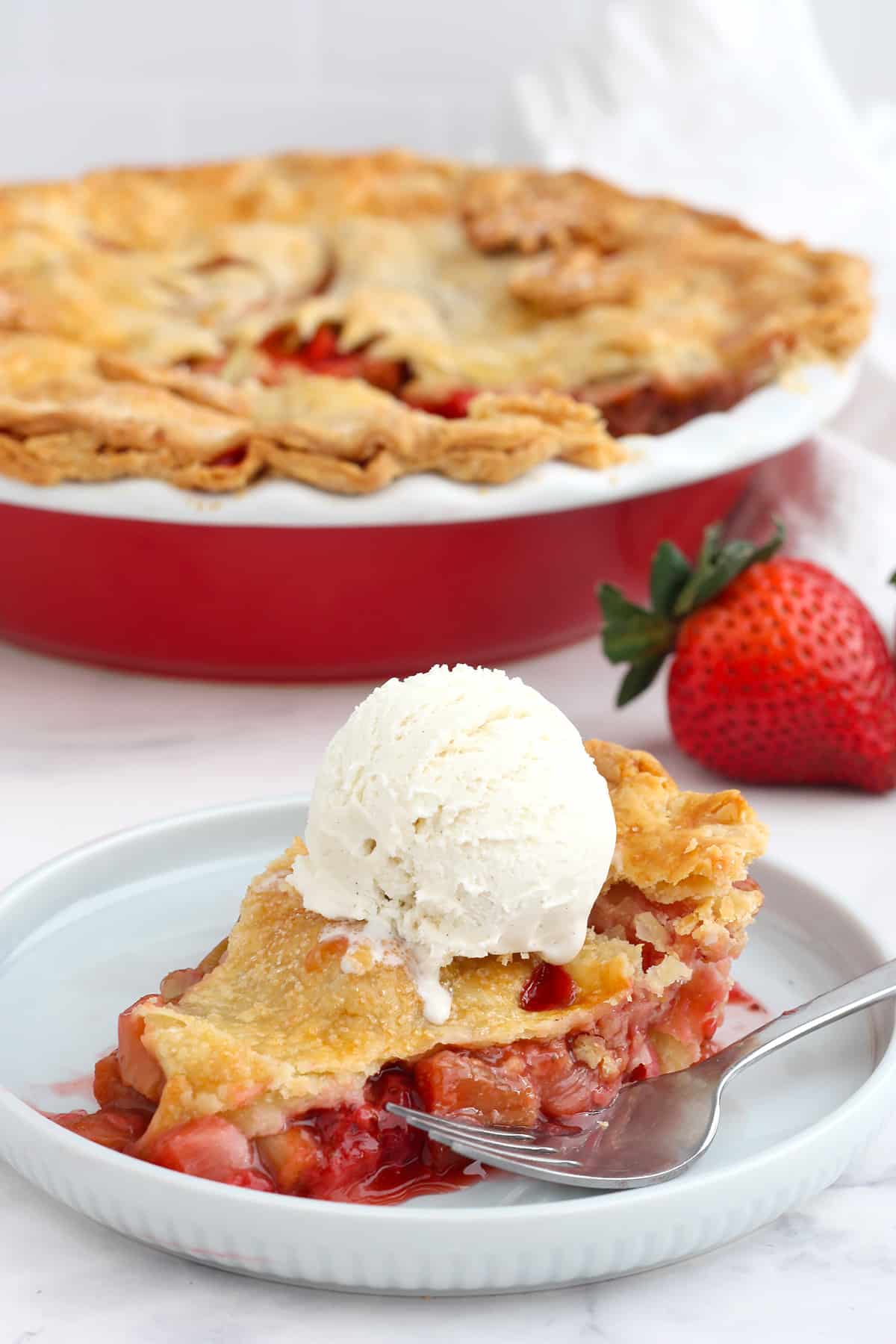 A slice of strawberry rhubarb pie topped with vanilla ice cream on a pale blue serving plate. 