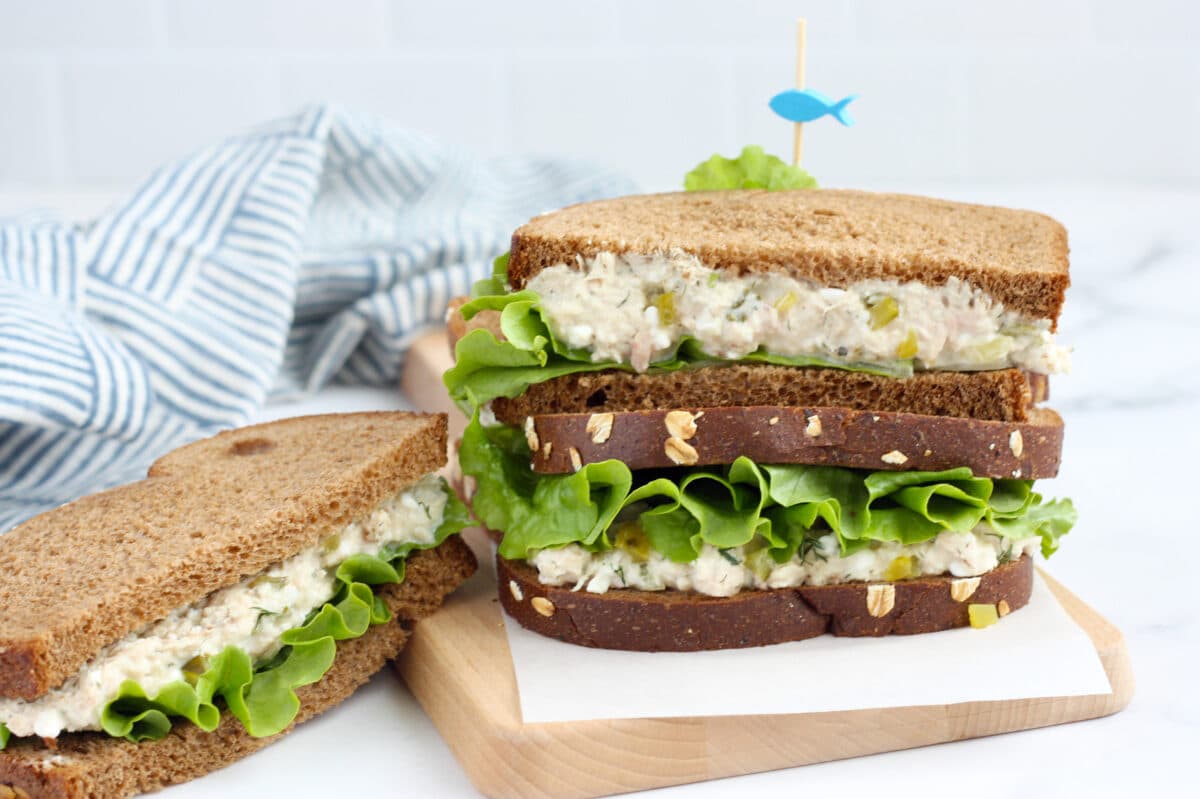 A tuna sandwich with lettuce on wheat bread stacked on a parchment lined cutting board with a toothpick. 