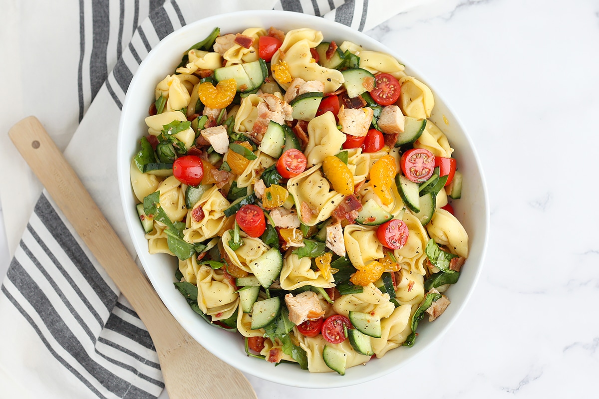 A large serving bowl filled with tortellini pasta salad on a striped linen with a wooden spoon.