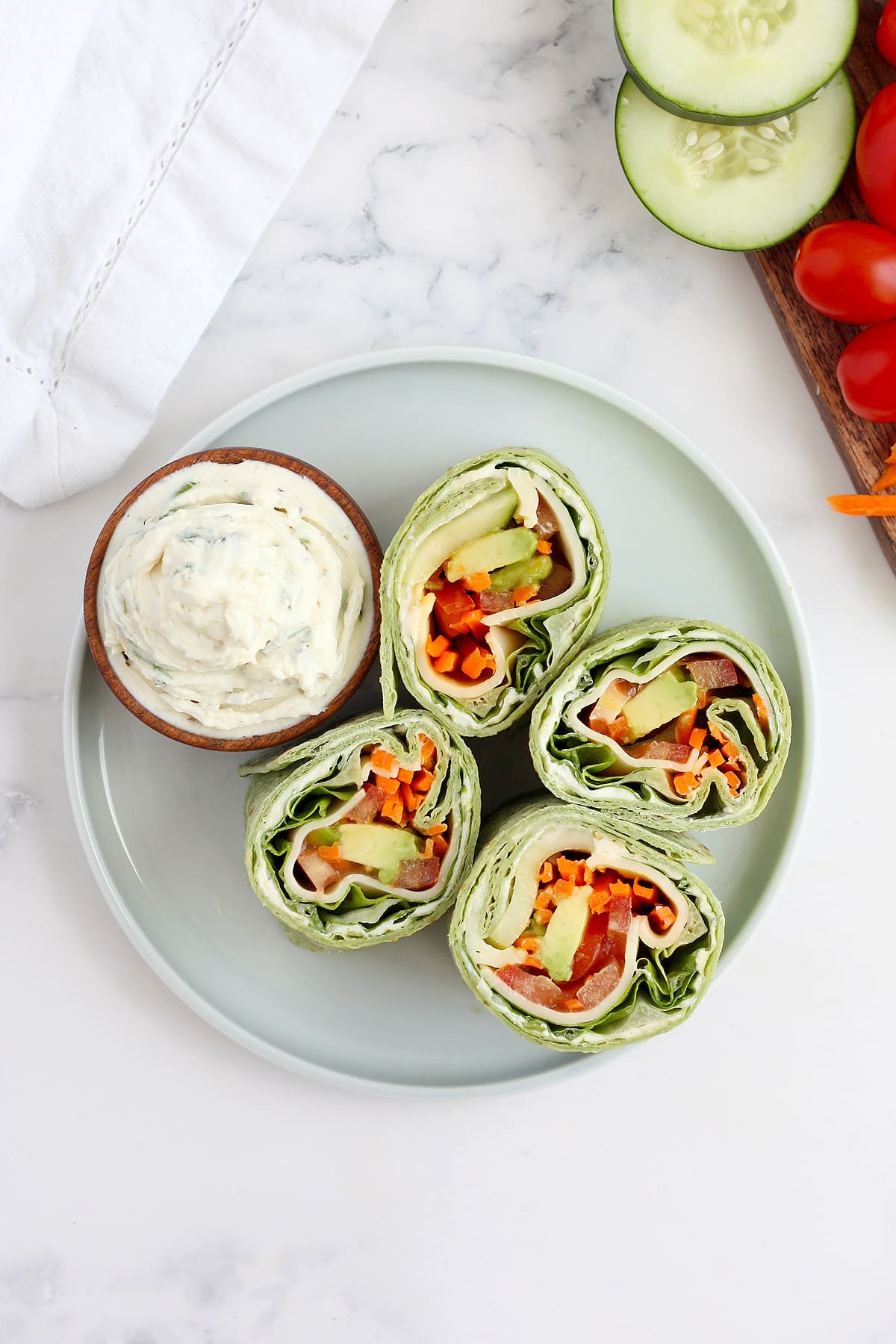 Veggie wraps cut into pinwheels on a blue serving plate with a bowl of herbed cream cheese. 
