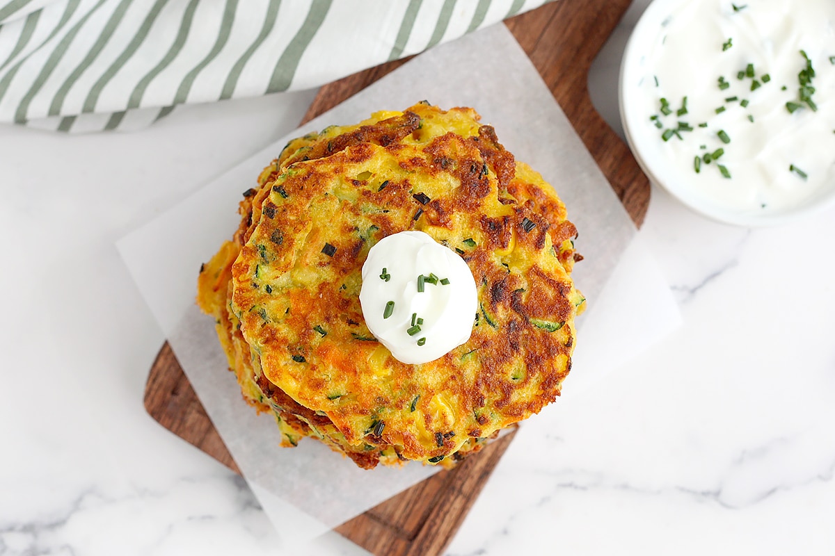 An overhead shot of vegetable fritters with a dollop of sour cream and chopped chives on a wooden cutting board. 